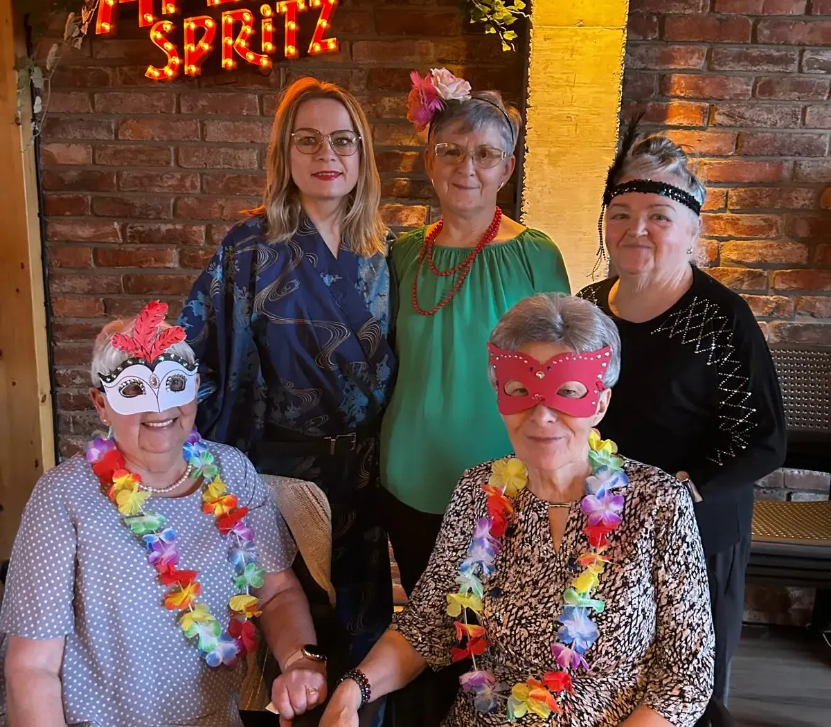 Grupa kobiet pozuje do zdjęcia podczas tematycznego spotkania, część z nich ma kolorowe maski i naszyjniki z kwiatów